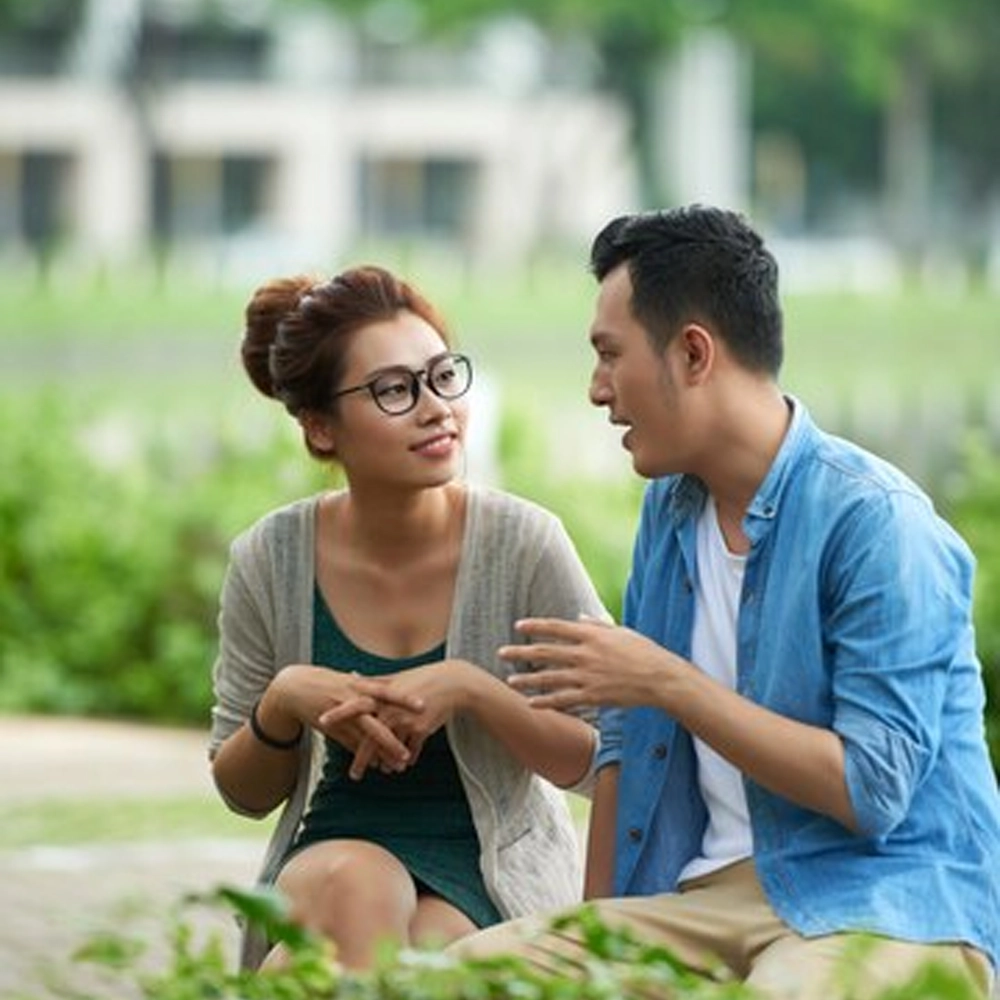 A young couple having a conversation in a park, sitting on a bench surrounded by greenery.