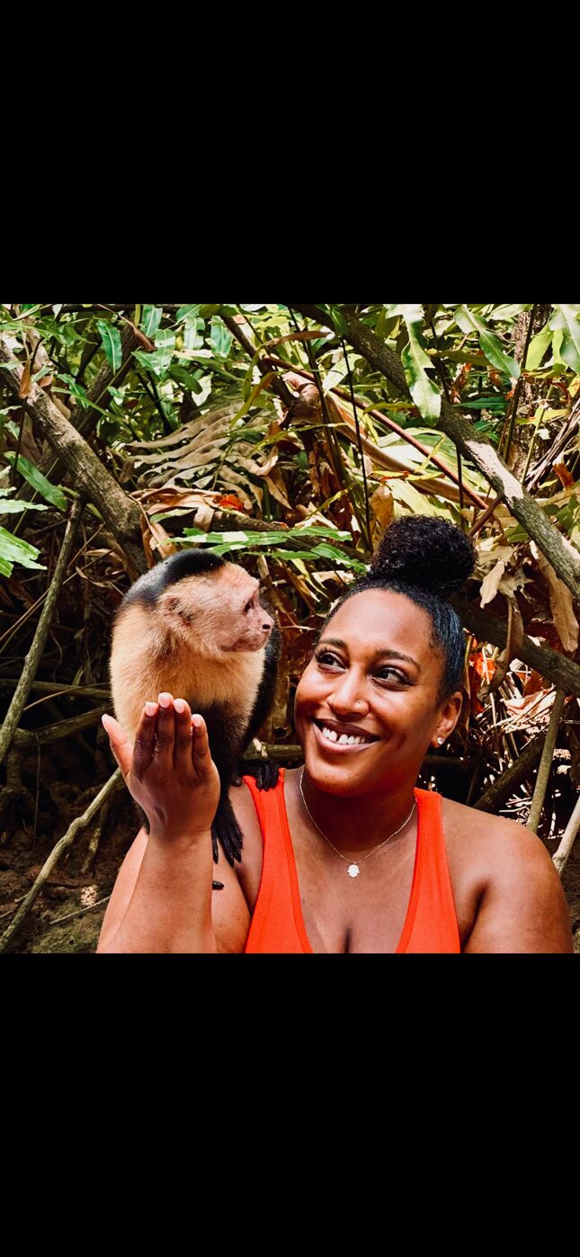 A smiling woman holding a small capuchin monkey in a lush, green outdoor setting.