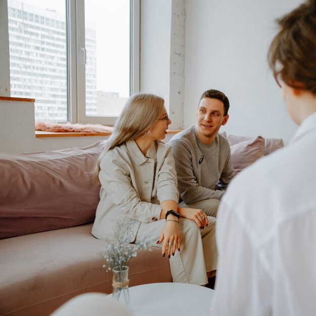 A young couple sitting on a pink couch, engaged in conversation with a third person.