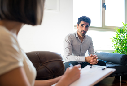 A man in a therapy session, sitting on a couch and looking at a therapist taking notes.